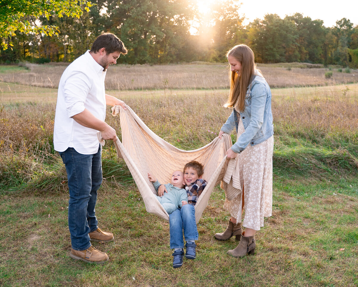 family playing in park swinging children while they are laying on blanket with sunset in background