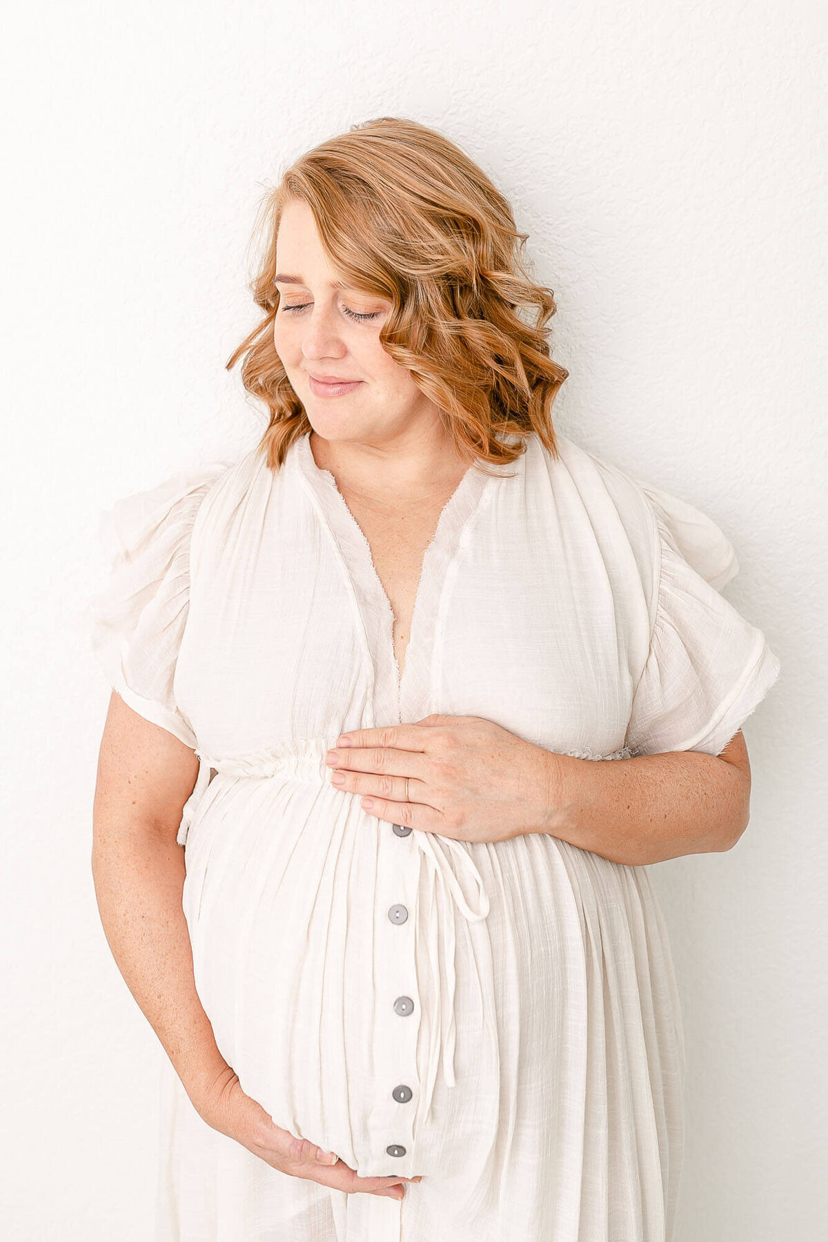 Woman with a baby bump standing in an all-white photography studio. She is wearing a long cream colored dress. She is looking down and has a soft smile on her face.