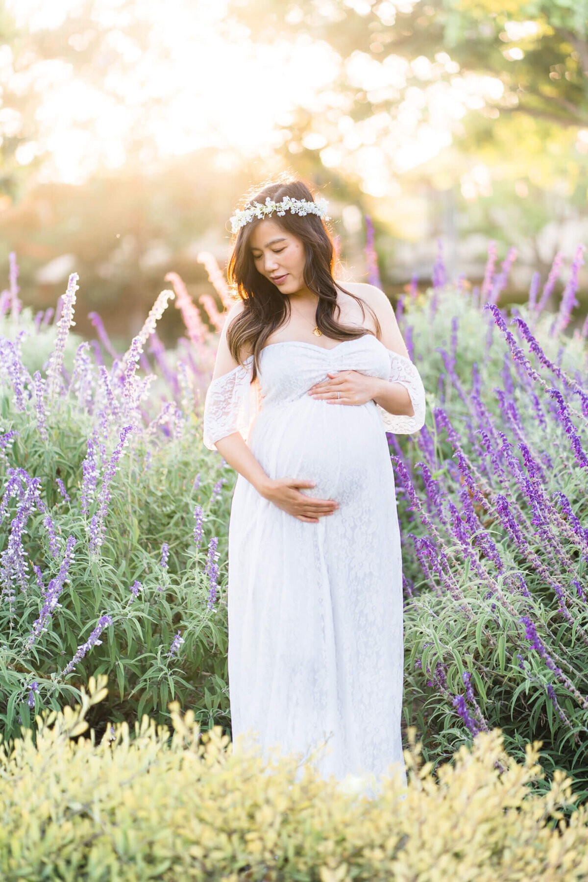 Pregnant mom in white dress holding her belly in purple field of flowers with sun in the background
