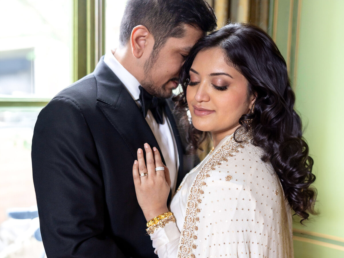 A couple lovingly embraces by a window. The man is wearing a black suit, and the woman is in a white embellished saree. They both have dark hair, and the setting appears warm and intimate.