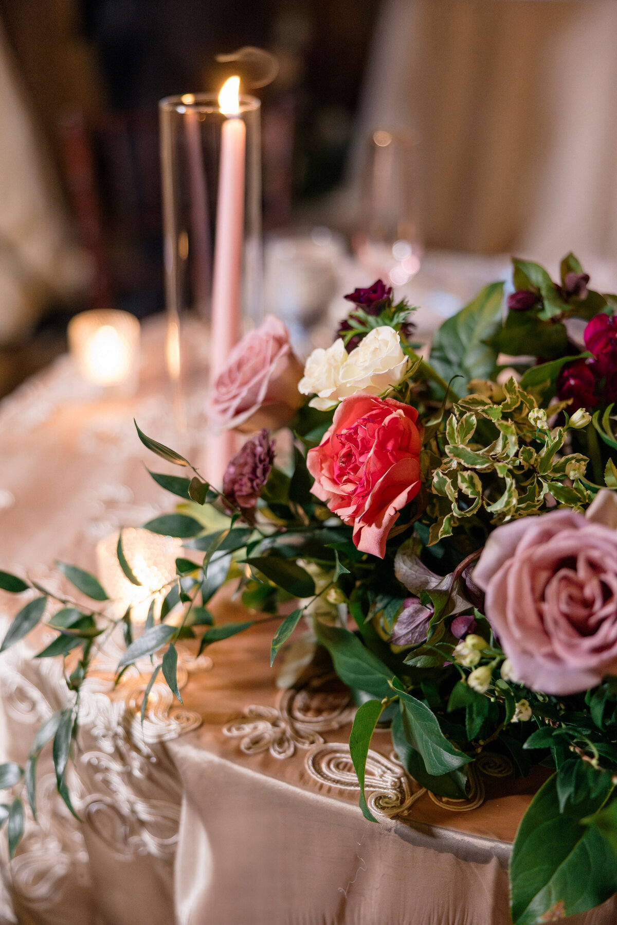 A decorative table setting featuring a lush floral arrangement with pink, white, and burgundy roses, green leaves, and a lit candle in a tall glass holder on an intricately embroidered tablecloth.