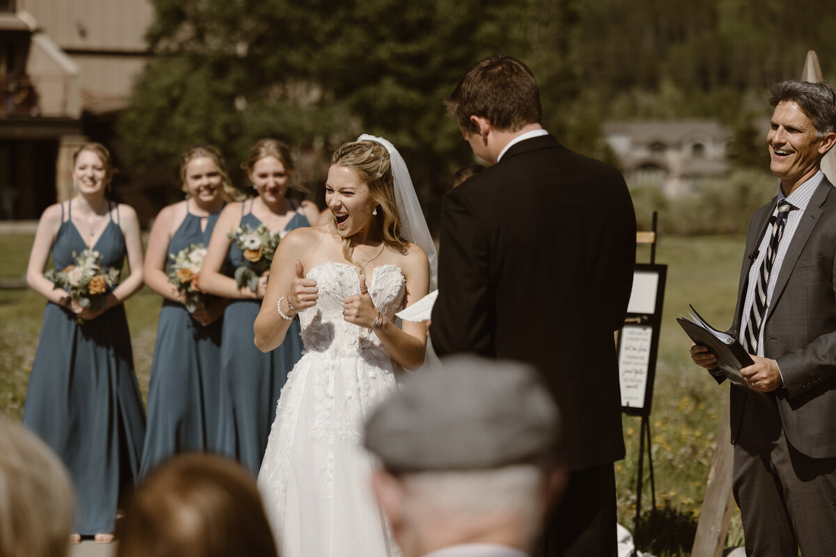 Bride gives two enthusiastic thumbs up during an outdoor wedding ceremony in Breckenridge, Colorado, surrounded by smiling bridesmaids and guests under the mountain sun.