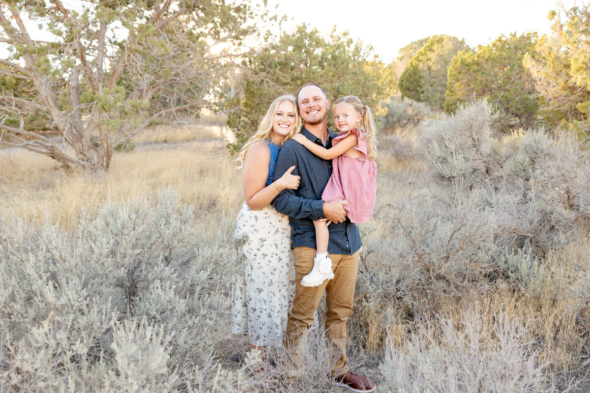 A father holds his daughter in his arms while the mother stands beside them, all smiling. The surrounding landscape of sagebrush and trees creates a picturesque scene.
