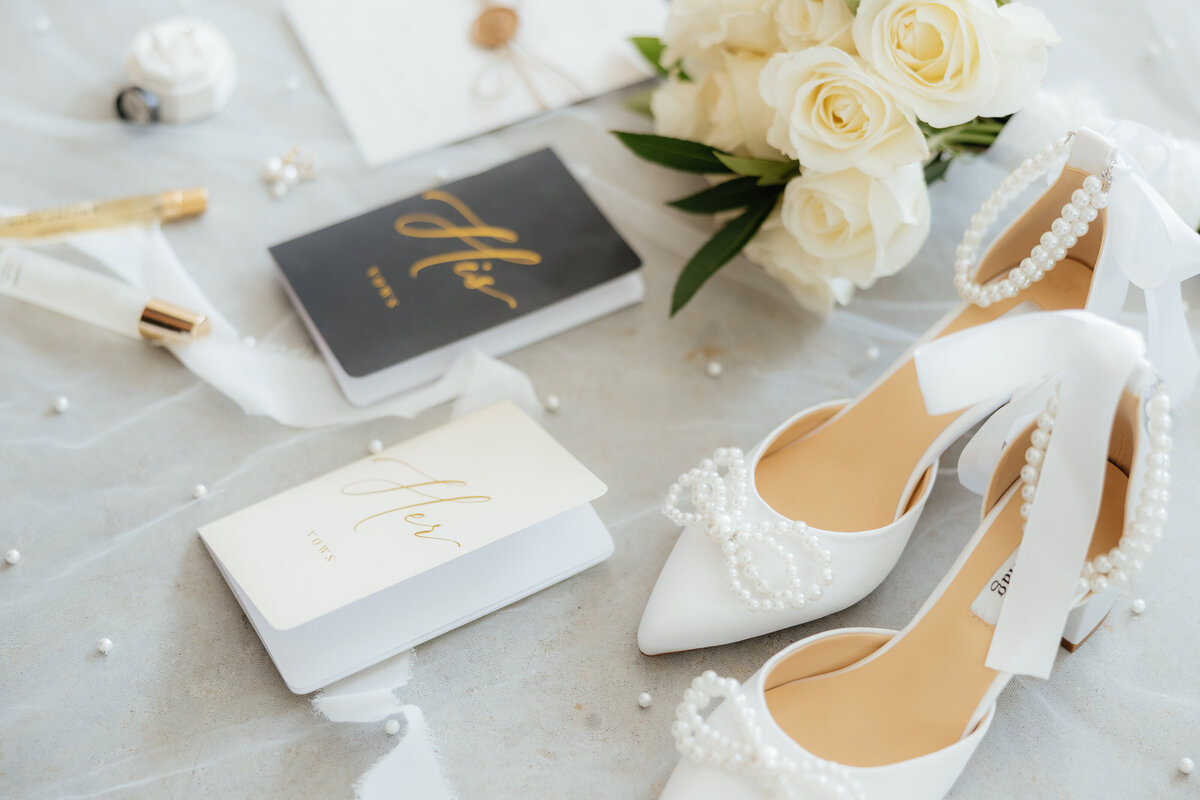 A detailed wedding flatlay with shoes, vow books, rings, perfume, florals, all laid out on a pearl veil.
