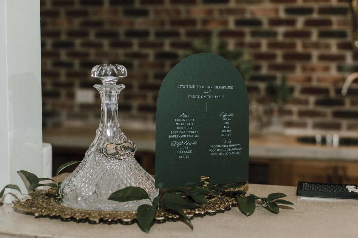 Rounded dark green place card with white cursive font on a gold place card holder atop gold table charger with glass bottle.