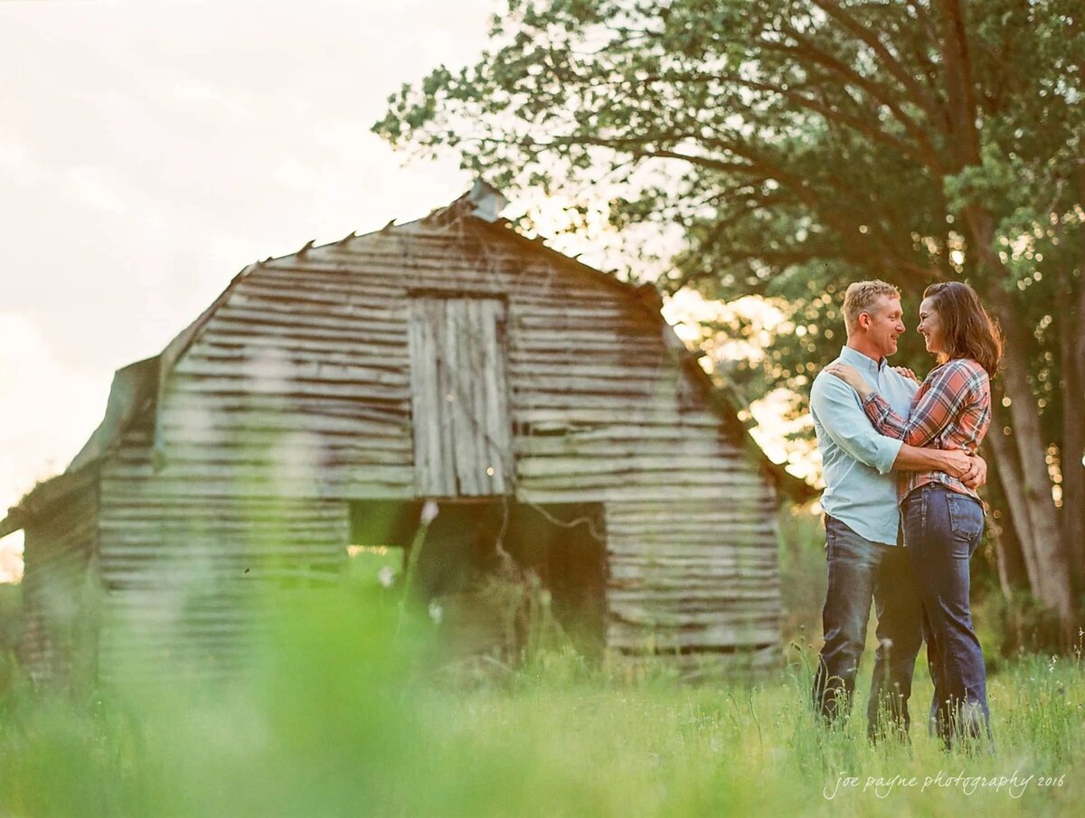 A couple standing in a rural field at dusk, with an old rustic barn in the background,