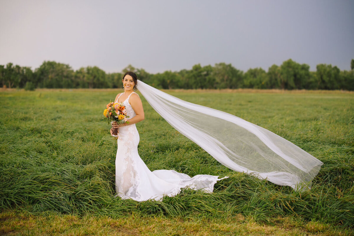 Bride standing alone in a green field with a flowing veil and bouquet