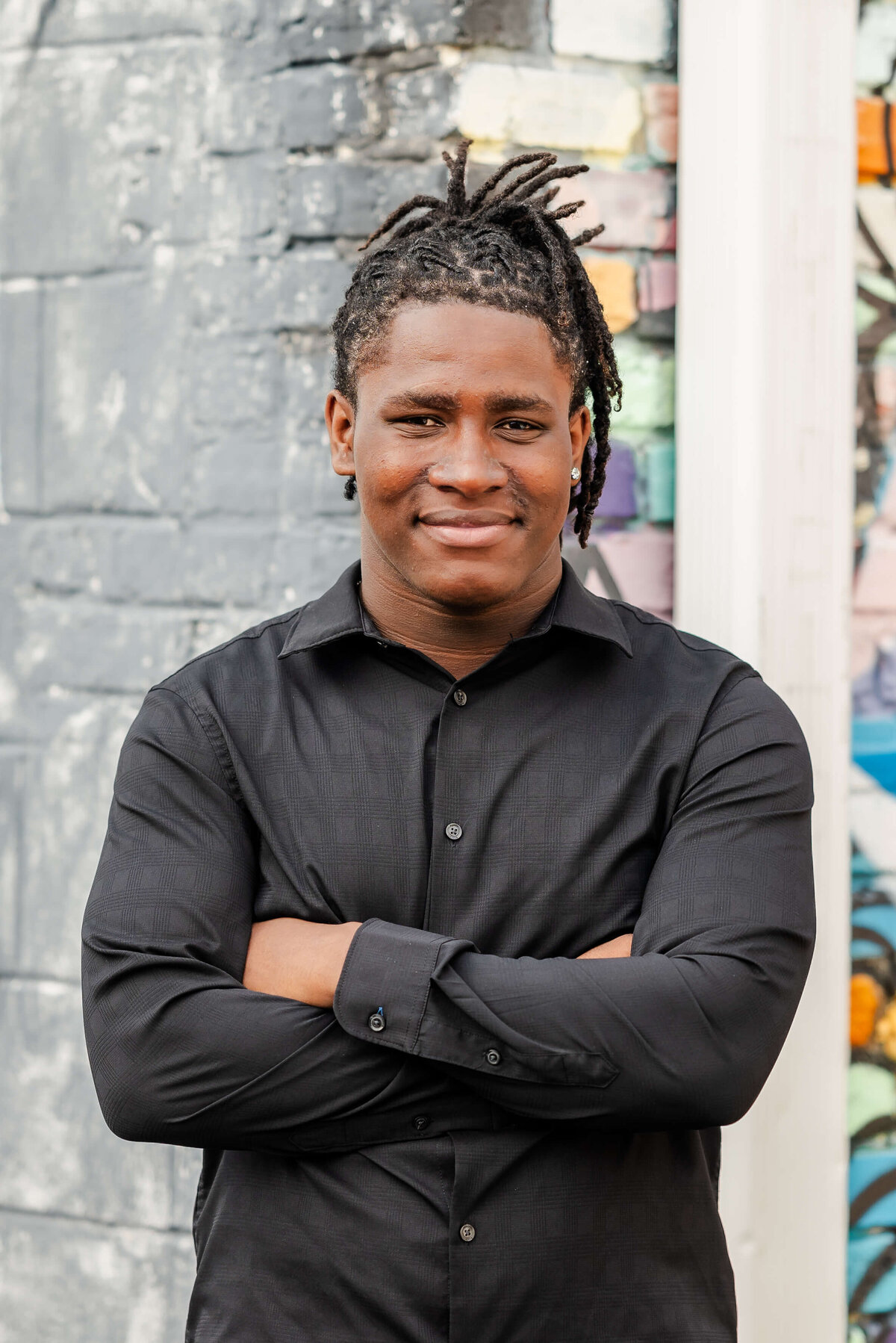 A high school senior, wearing a black button down, crosses his arms and smiles. Behind him you can see a colorful mural.