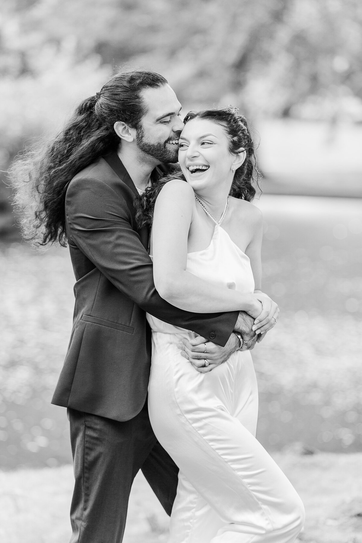 A woman stands with her back to a man at their Boston Common engagement photoshoot. The man's arms are wrapped around the woman's waist and her hands overtop of his. His nose is resting against her temple and they are both laughing. Captured by New England Wedding Photographer Lia Rose Weddings