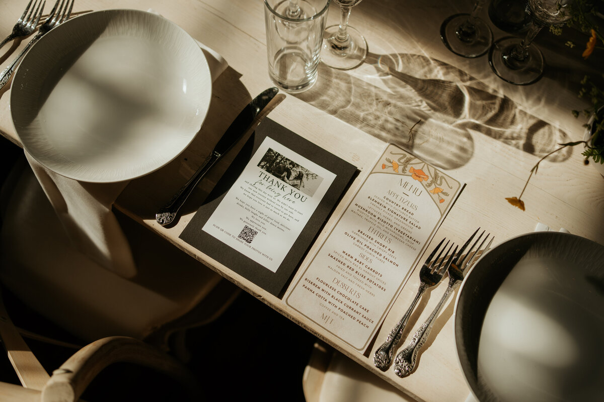 Close-up of a beautifully set wedding reception table with a white plate, silver cutlery, and a detailed menu card. Next to the menu is a thank you note with a black and white photo of the couple. The table is illuminated by natural light, creating moody shadows.