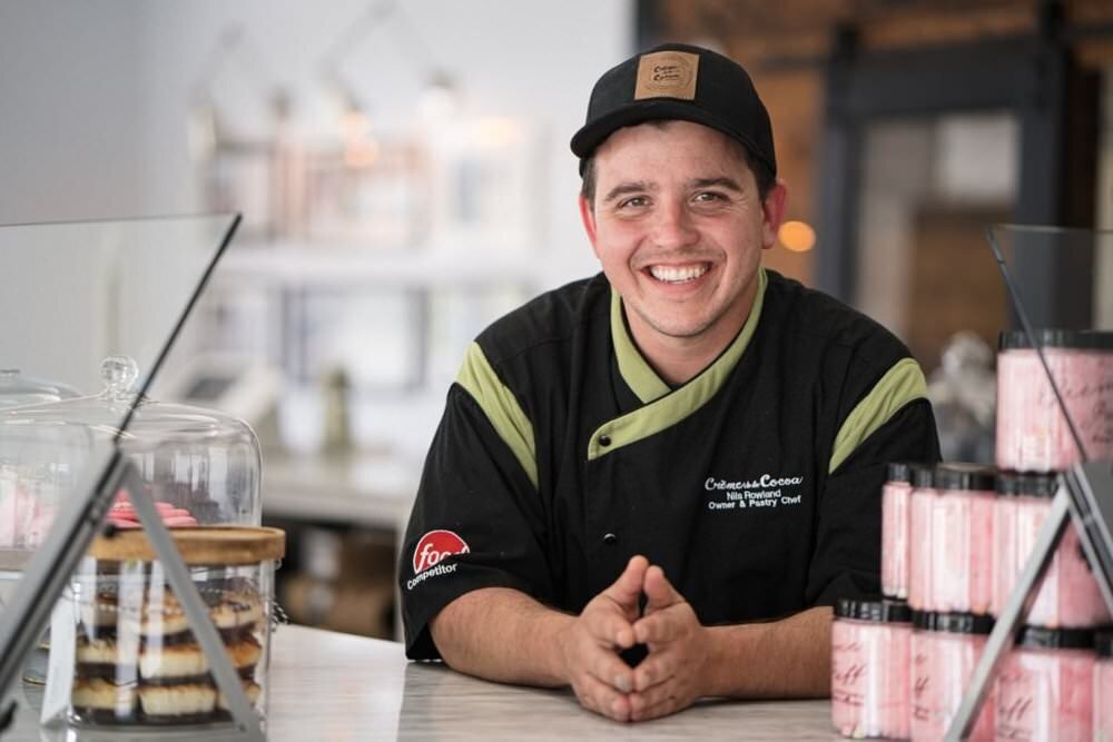 Smiling-pastry-chef-in-his-bakery-wearing-a-black-and-green-uniform-surrounded-by-delicious-desserts-and-cakes-on-display