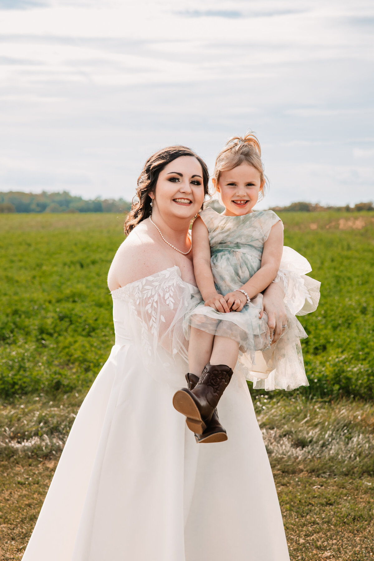 A bride holds her young flowergirl in. her arms while smiling in a field