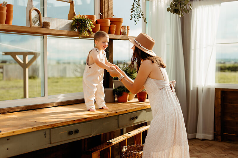 Mom holds hands of toddler standing on a a work bench in a greenhouse while eating a white dress and fashionable hat