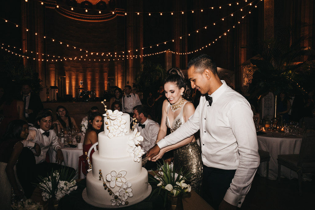 The stunning photo captures a couple in formal attire cutting a tiered wedding cake adorned with white flowers at a reception in Washington D.C.'s elegant Mellon Auditorium. The scene exudes a romantic ambiance, illuminated by soft string lights, with guests gathered around the table, some seated and some standing, witnessing the special moment. The venue, known for its luxurious European flair, adds to the opulence of the event, making it a truly unforgettable experience captured beautifully by photographer Sarah Bradshaw.