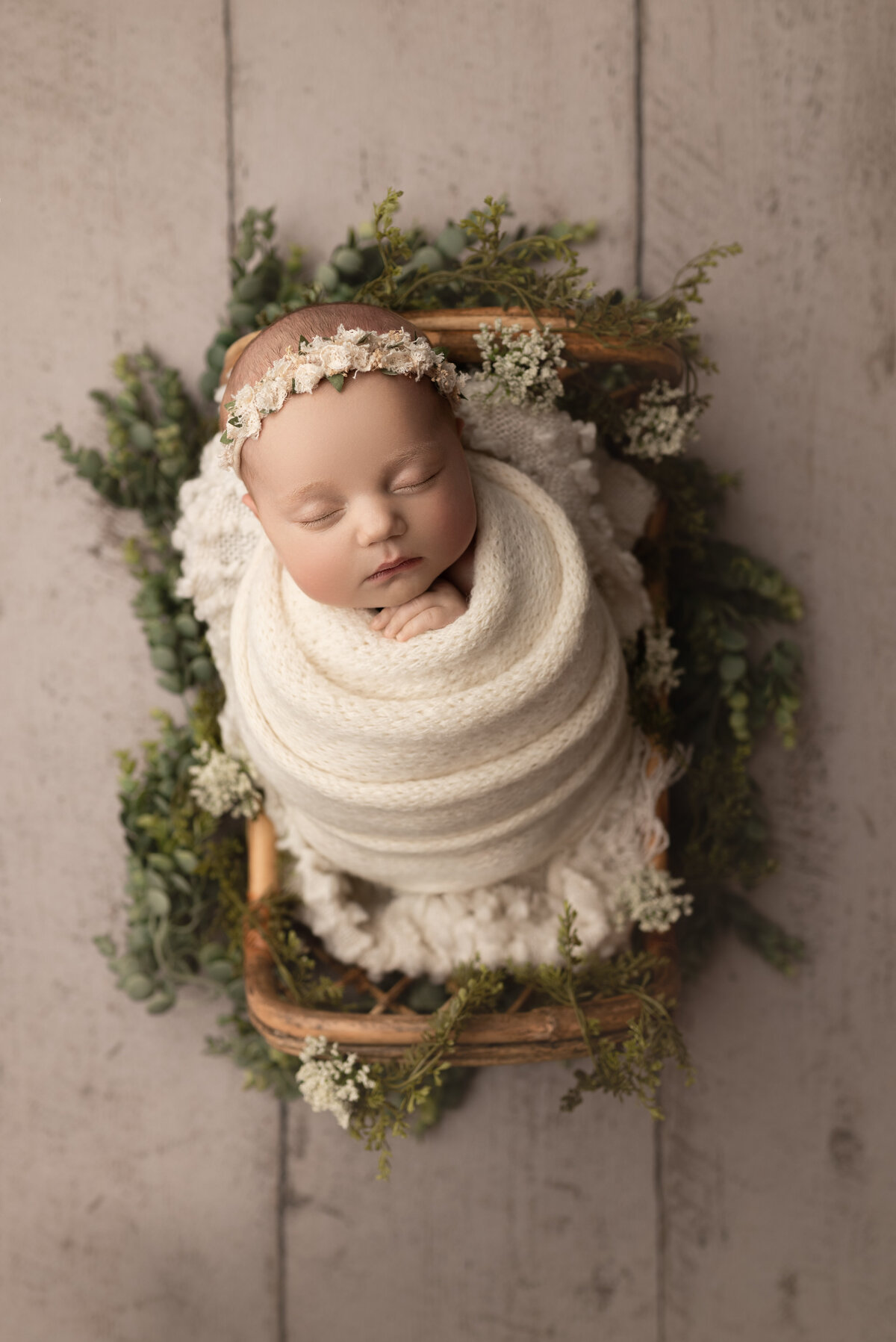 Fine art newborn photos captured by best Main Line newborn photographer Katie Marshall. Baby girl is swaddled in a cream knit swaddle and matching delicate floral headband. She is placed in a wood basket adorned with soft greenery.