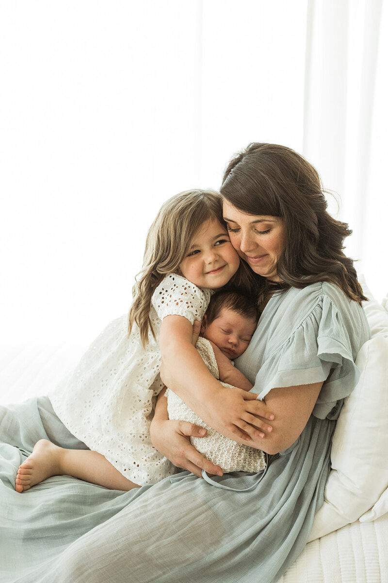 Mom sits on bed holding and hugging her newborn daughter & toddler daughter