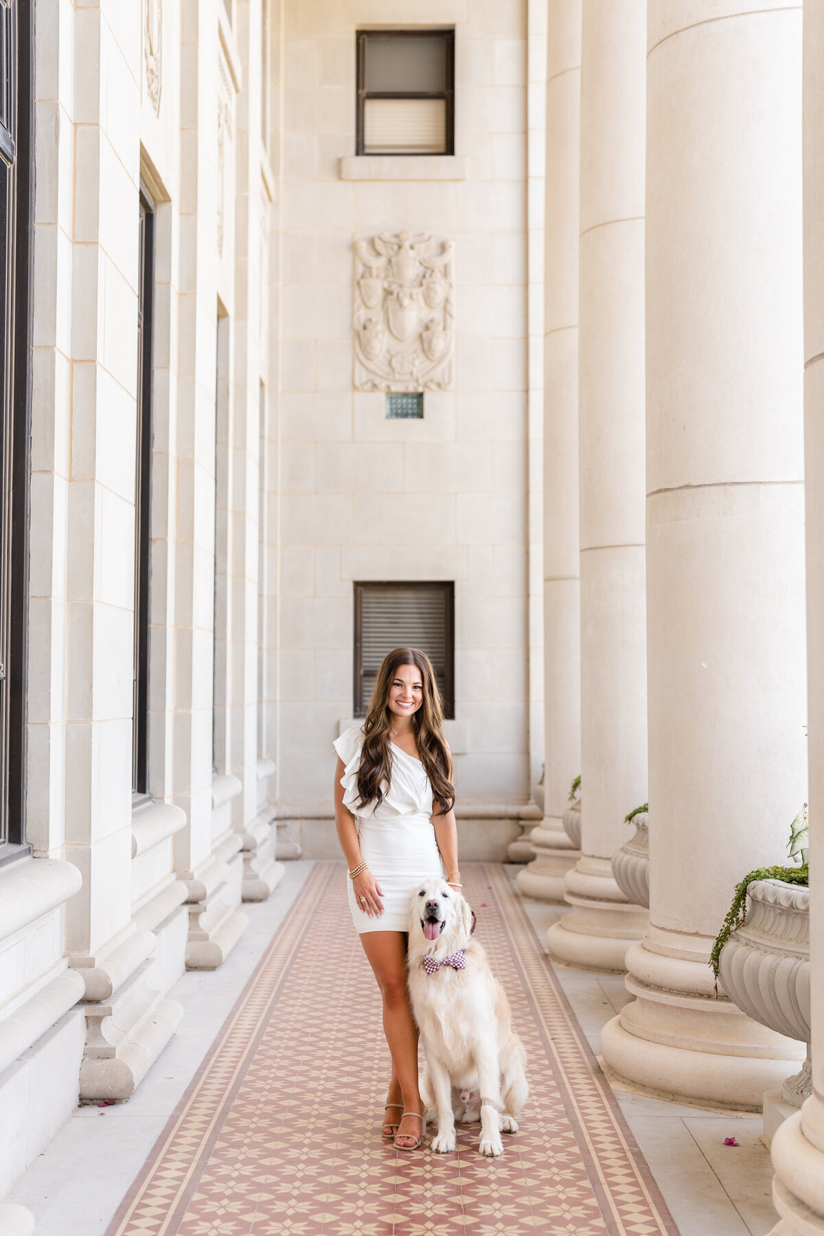 Texas A&M senior girl standing in white dress with her dog by her side and smiling in the columns of the Administration Building