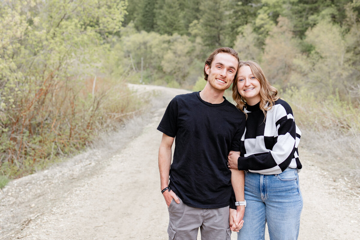 A couple standing close together on a scenic dirt road in Provo Canyon, smiling at the camera with vibrant trees in the background.