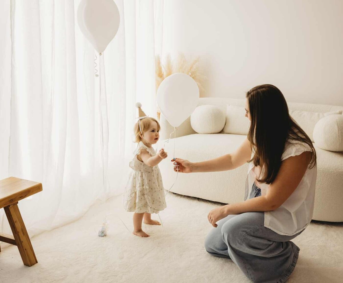Ruua Hamdan Photography (UK): A toddler in a party hat and floral dress stands on a white rug, reaching for a white balloon held by a kneeling adult.