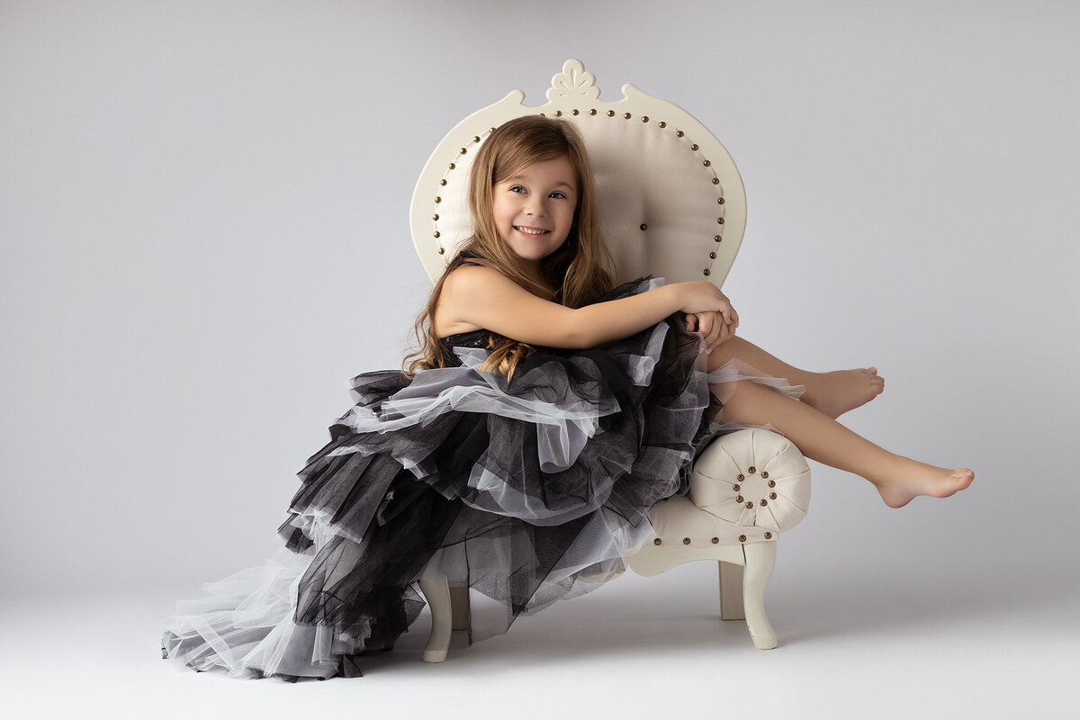 Lindsay Walden Photography captures a young girl in a black and white ruffled dress, sitting gracefully on a vintage chair.