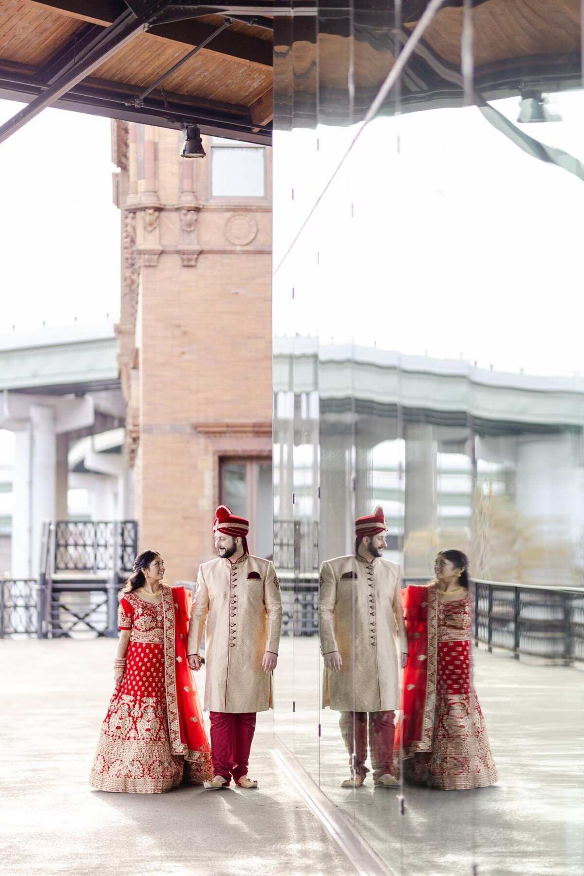A couple in traditional south asian wedding attire standing under a covered pavilion, reflected in a vertical mirror.