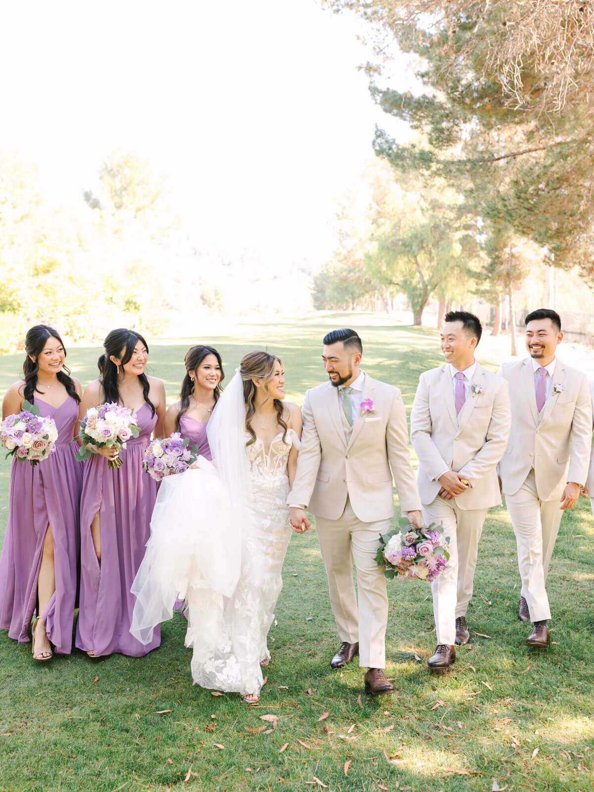 A joyful wedding party strolls on a sunny lawn. The bride in a white gown holds hands with the groom in a beige suit. Bridesmaids in lilac dresses and groomsmen in matching suits smile, holding bouquets. Trees and grass create a serene backdrop.