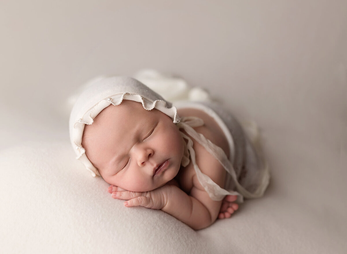 A sleeping newborn baby wears a white bonnet on a white pad in a studio