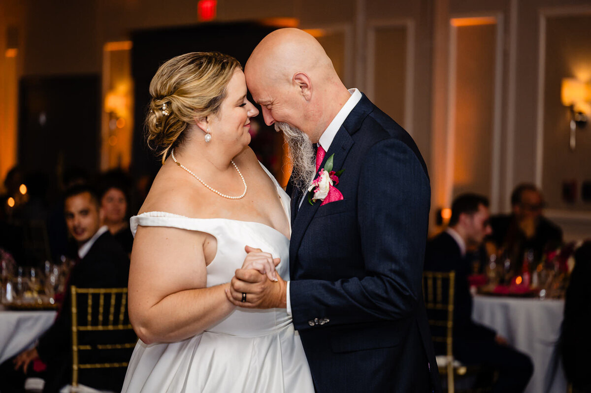 A bride and groom share an intimate dance, foreheads touching with tender expressions, in a ballroom setting