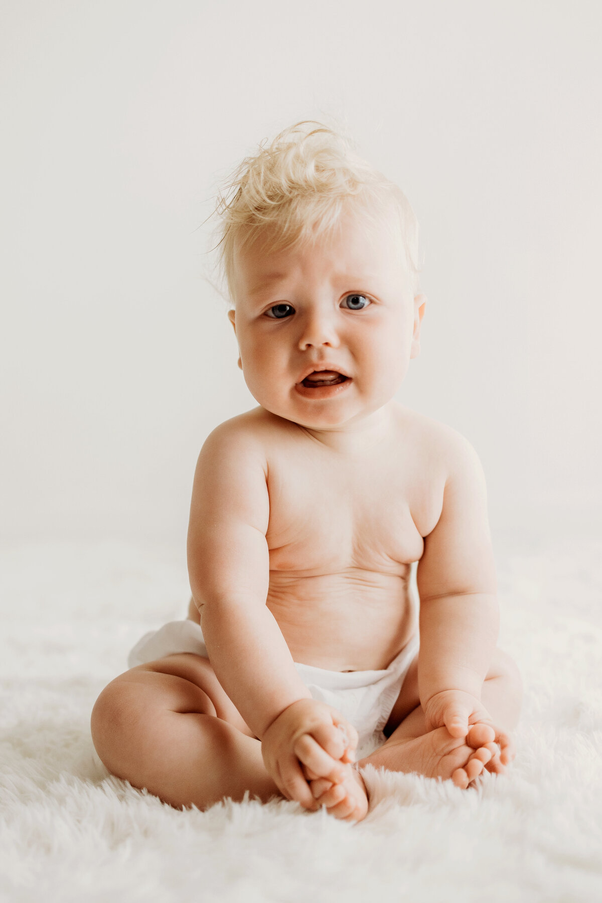 A pouty faced six month old baby boy sits on a white rug with a white background with only a diaper cover on for his Denver milestone session.