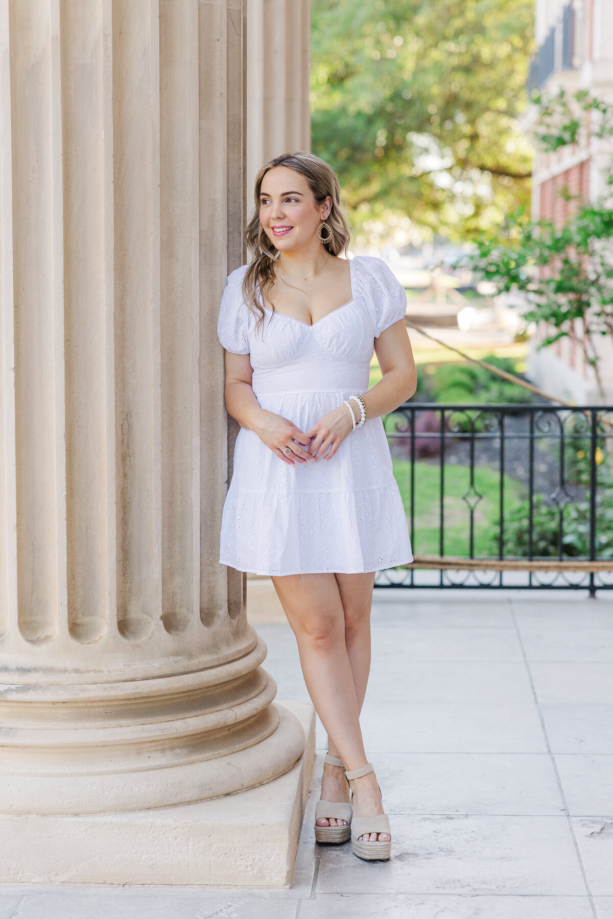 High school senior wearing a white eyelet dress and platform heels leans against a tall classical column with a soft smile. A sunny and elegant portrait featuring architectural details and greenery in the background.