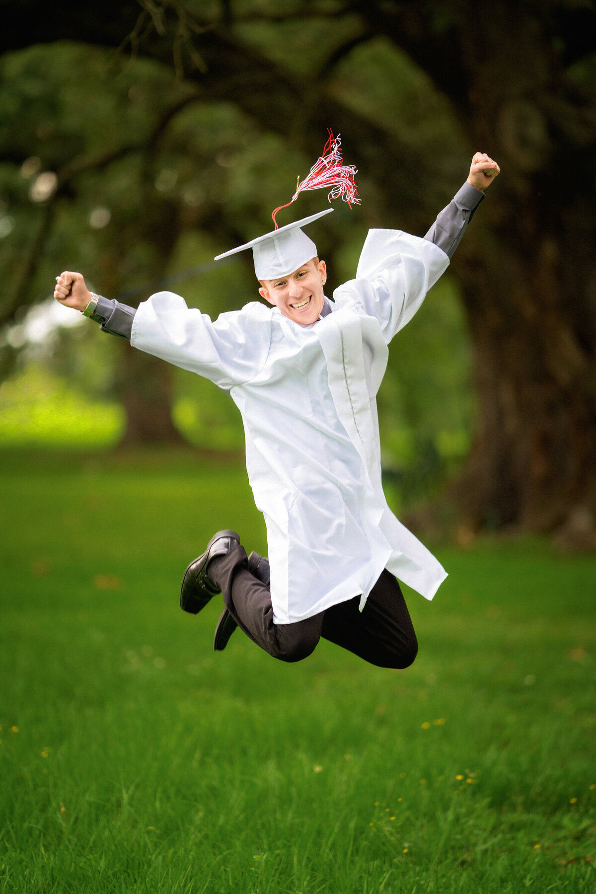 Senior boy wearing his white cap and gown and jumping in the air with excitement.  He is wearing black pants and shoes.  The tassel of his cap is flying up.  His arms are up in the air.  There are oak trees and grass in the background.