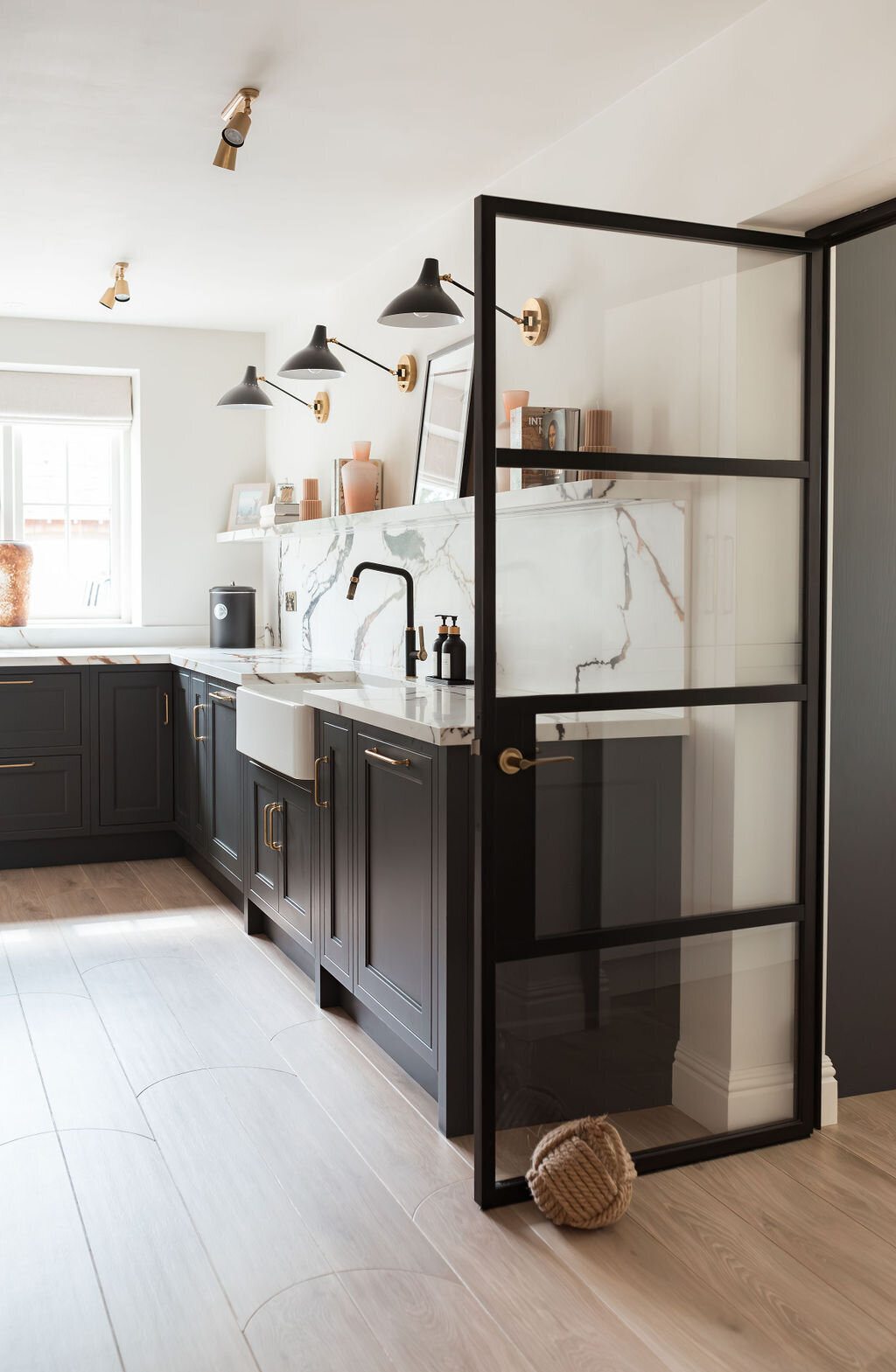 A modern kitchen with dark lower cabinets, a white marble backsplash with black and gold veining, a farmhouse sink, and minimalist open shelving holding books and decor. A glass door with black framing is propped open to the right by an oversized yarn ball doorstop.