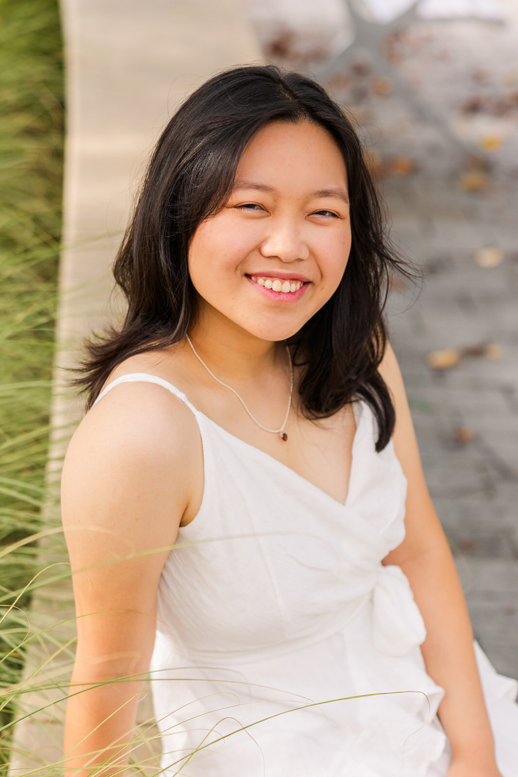 close up of high school girl sitting on a small wall looking sideway at the camera with grass in the foreground during senior portraits