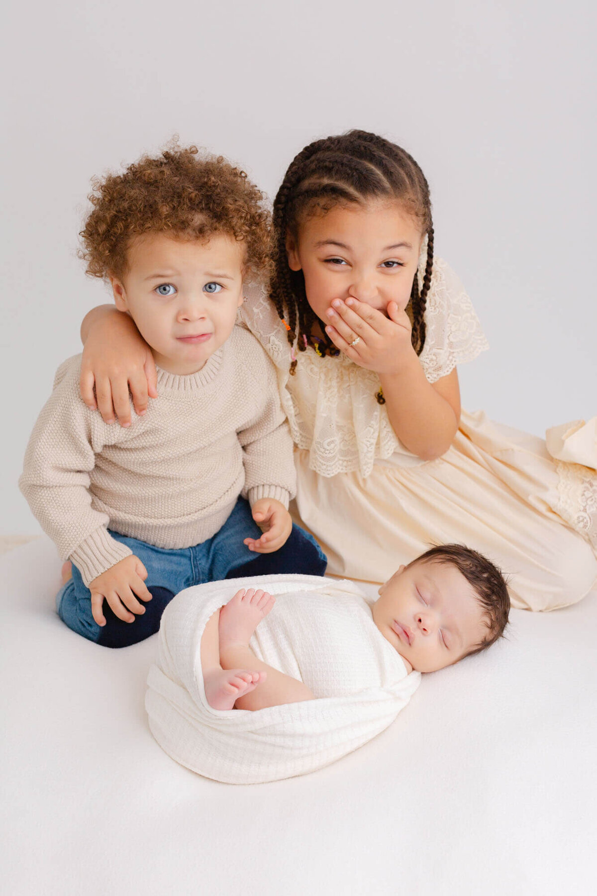 Big sis and toddler brother are sitting behind  newborn baby brother. Sister has her hand over her mouth like she's got secret she's laughing about. Cute photo of siblings with their new baby brother during his newborn photoshoot.