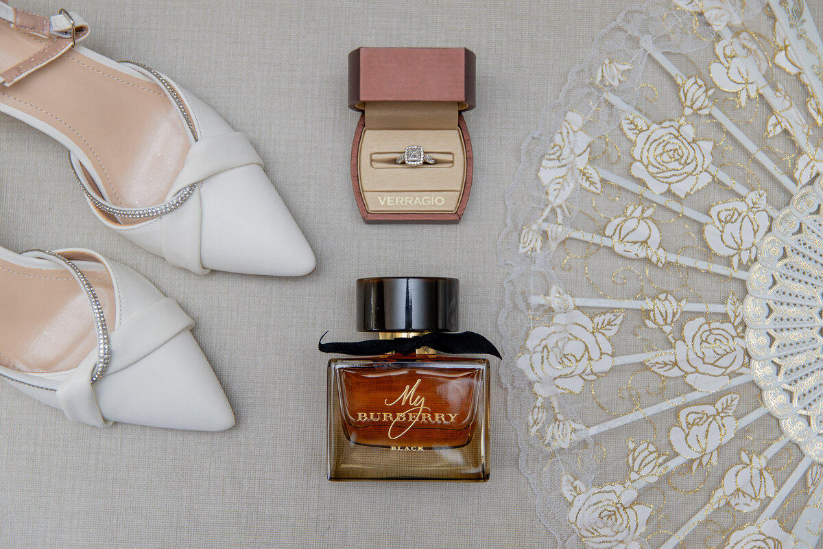 Flat lay of white wedding day shoes, cologne, a ring box with her wedding ring, and a lacy fan.