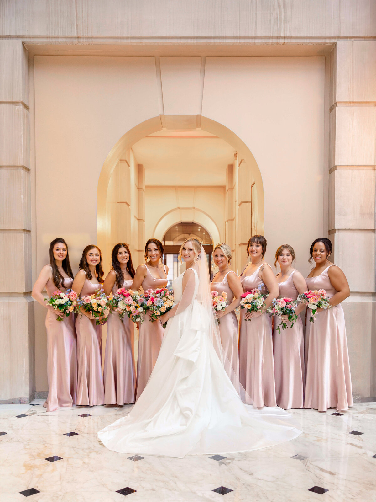 A bride in a white gown stands with her back to the camera, surrounded by eight bridesmaids in matching blush dresses. They hold colorful bouquets and stand in a grand hall with marble flooring and archways.