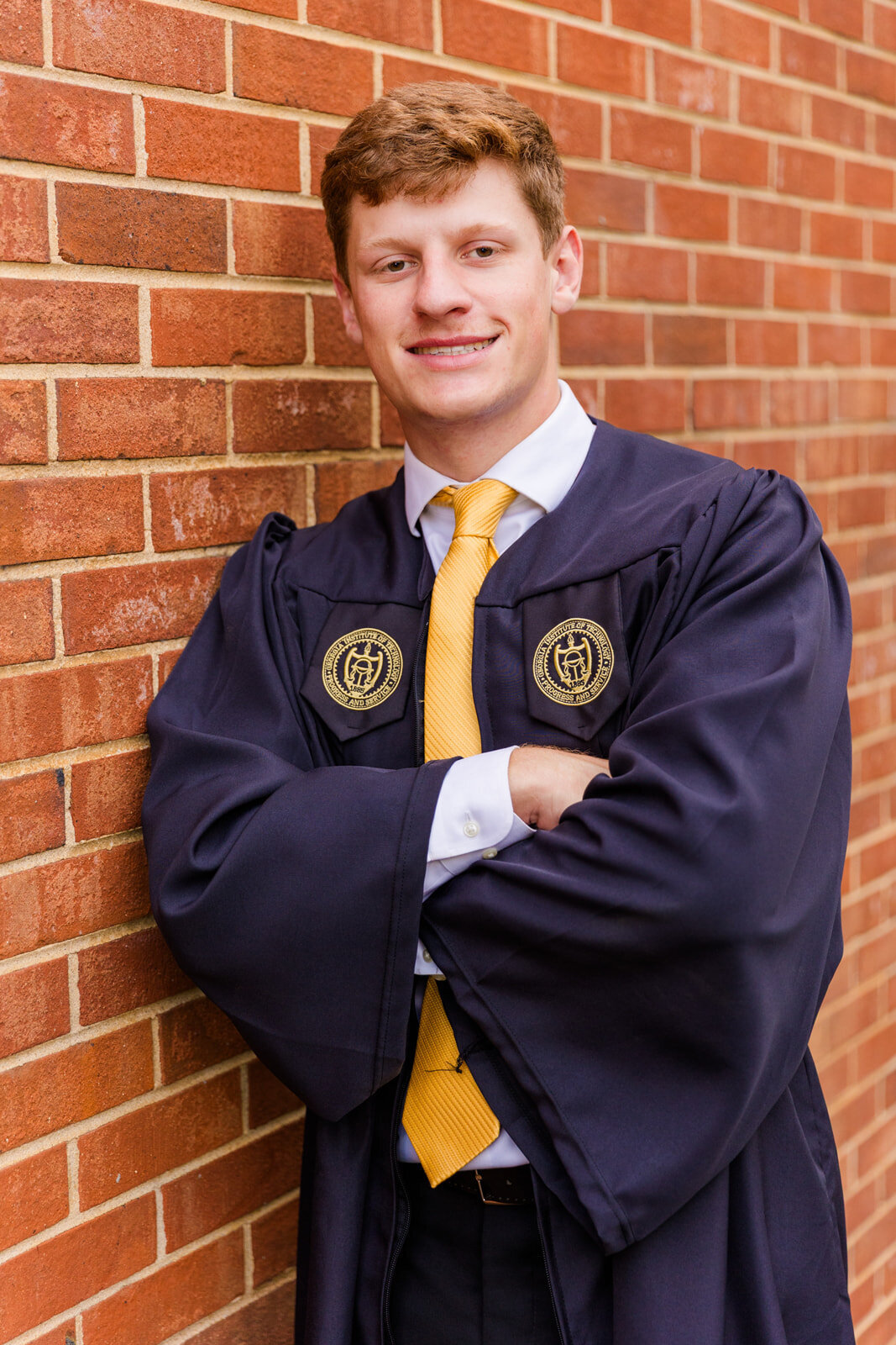 Georgia Tech University senior boy leaning against a red brick wall wearing cap and gown during graduation photoshoot on campus by Atlanta photographer Laure Photography