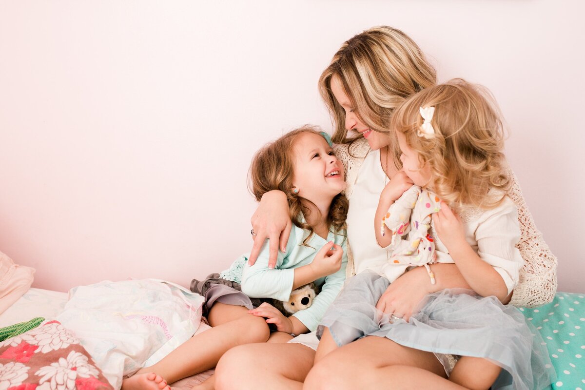 A mother embraces her two young daughters as they share a joyful moment together on a bed with pink bedding, captured by a talented Pittsburgh family photographer.
