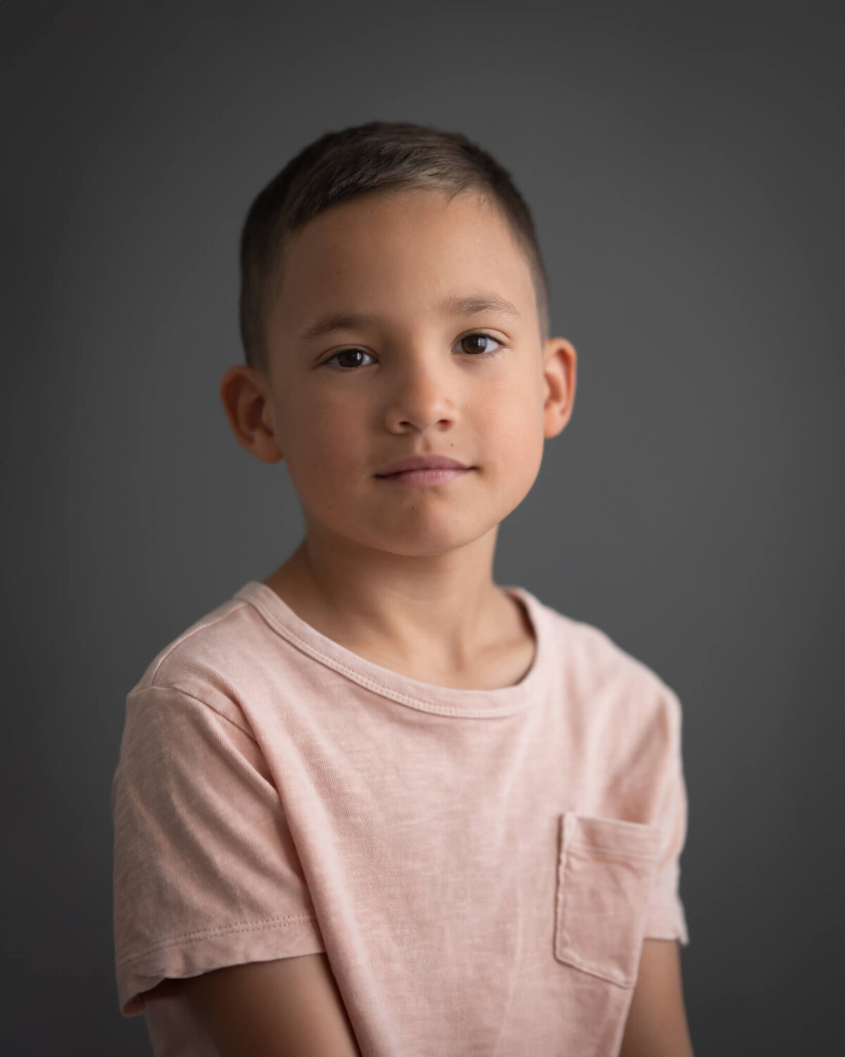 studio portrait of boy in light pink shirt with grey backdrop and a serious face