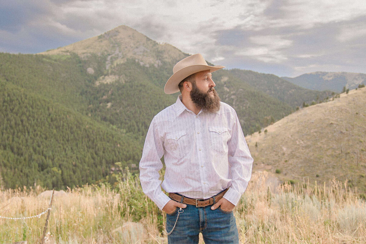 The groom, attired like a cowboy, looks out at the mountains surrounding the location of his Colorado wedding