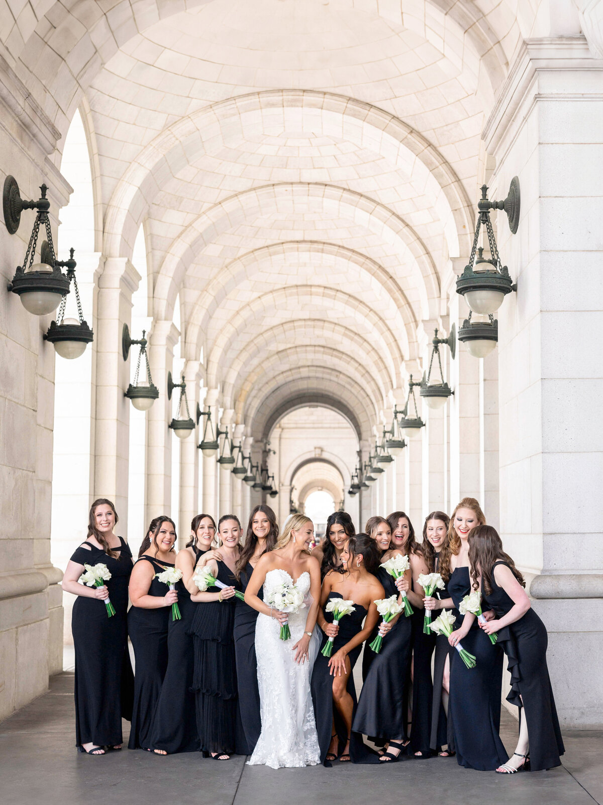 A bride in a white dress stands with her bridesmaids, who are wearing black dresses, under a series of arched hallways. They are all holding white bouquets and smiling at the camera. The background features elegant architectural details.