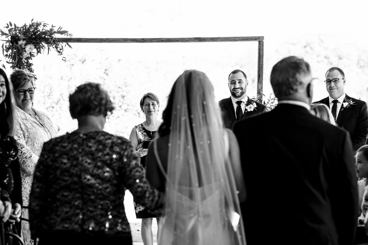 Black-and-white-image-taken-from-behind-the-bride-over-her-shoulder-as-she-is-being-walked-down-the-aisle-focused-on-her-smiling-groom-at-Upstairs-Atlanta