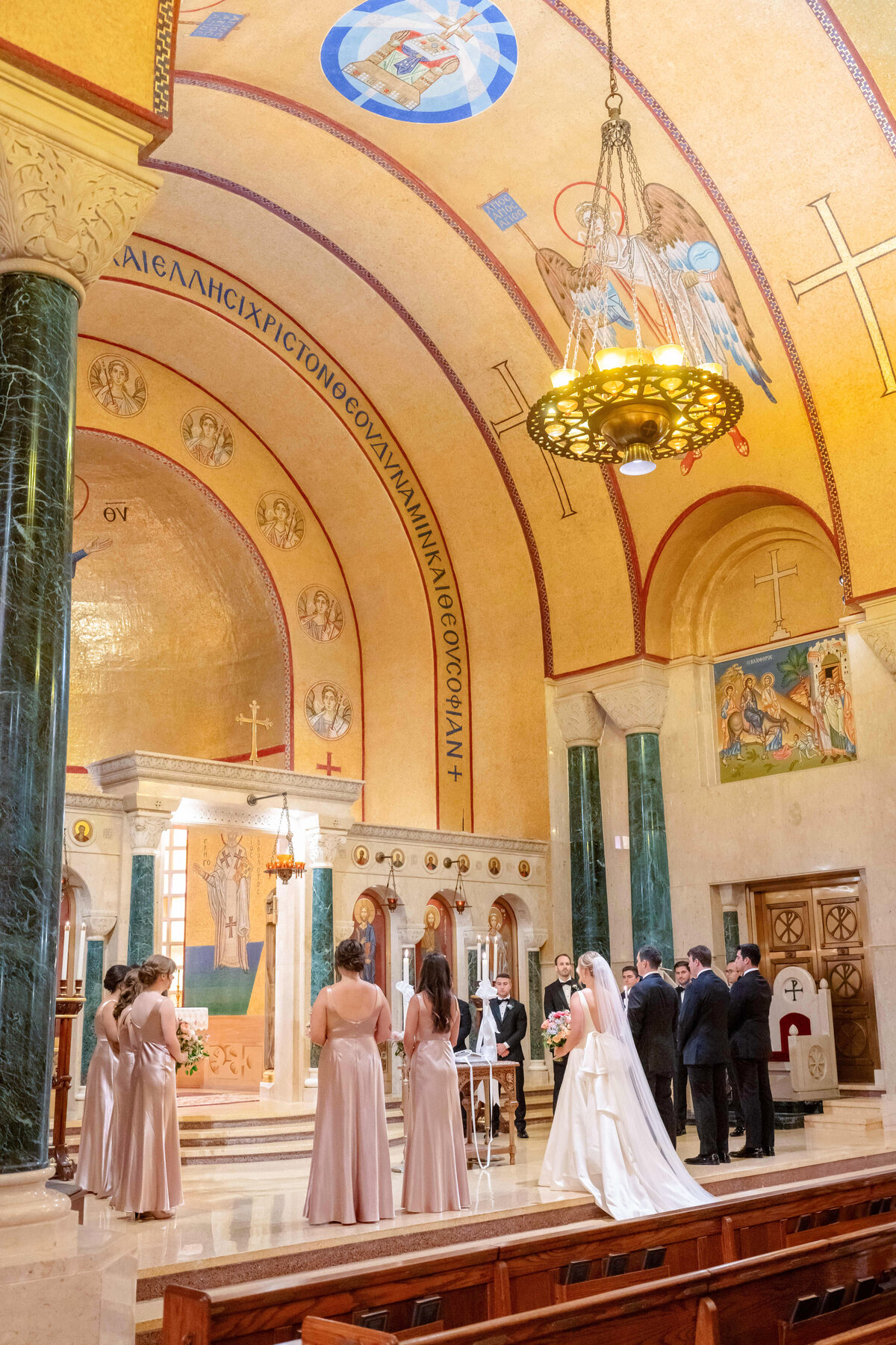 A wedding ceremony in a grand church with ornate architecture. The bride and groom stand at the altar, surrounded by bridesmaids in light pink dresses and groomsmen in dark suits. The church features decorative arches and a large chandelier.