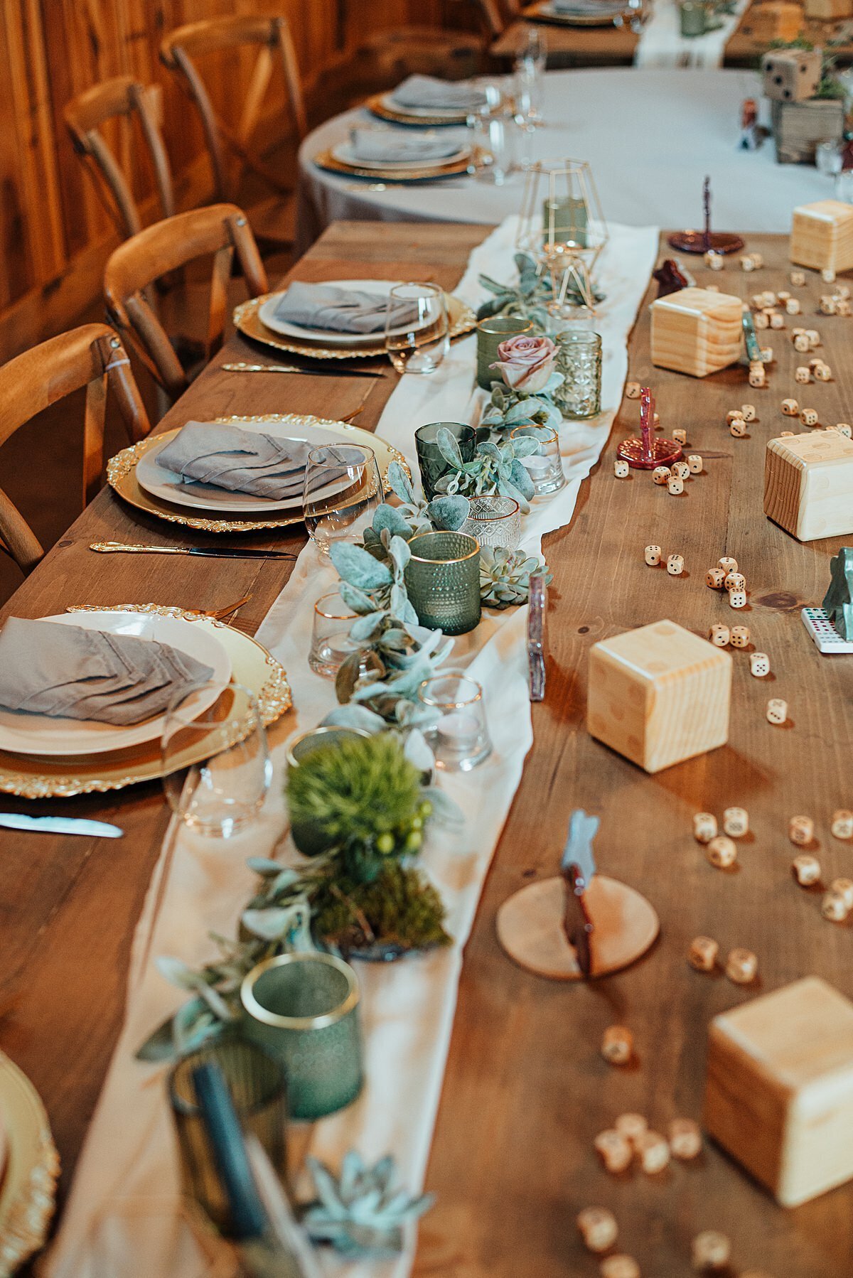Long barn wood farm tables set for the bridal party are set with ivory chargers, white plates and gray napkins. An ivory organza table runner runs down the center of the tables. The table runner is decorated with lambs ear, green and gold votive candles and succulents. The front of the table is decorated with dice, over sized dice and meeples for a board game theme.