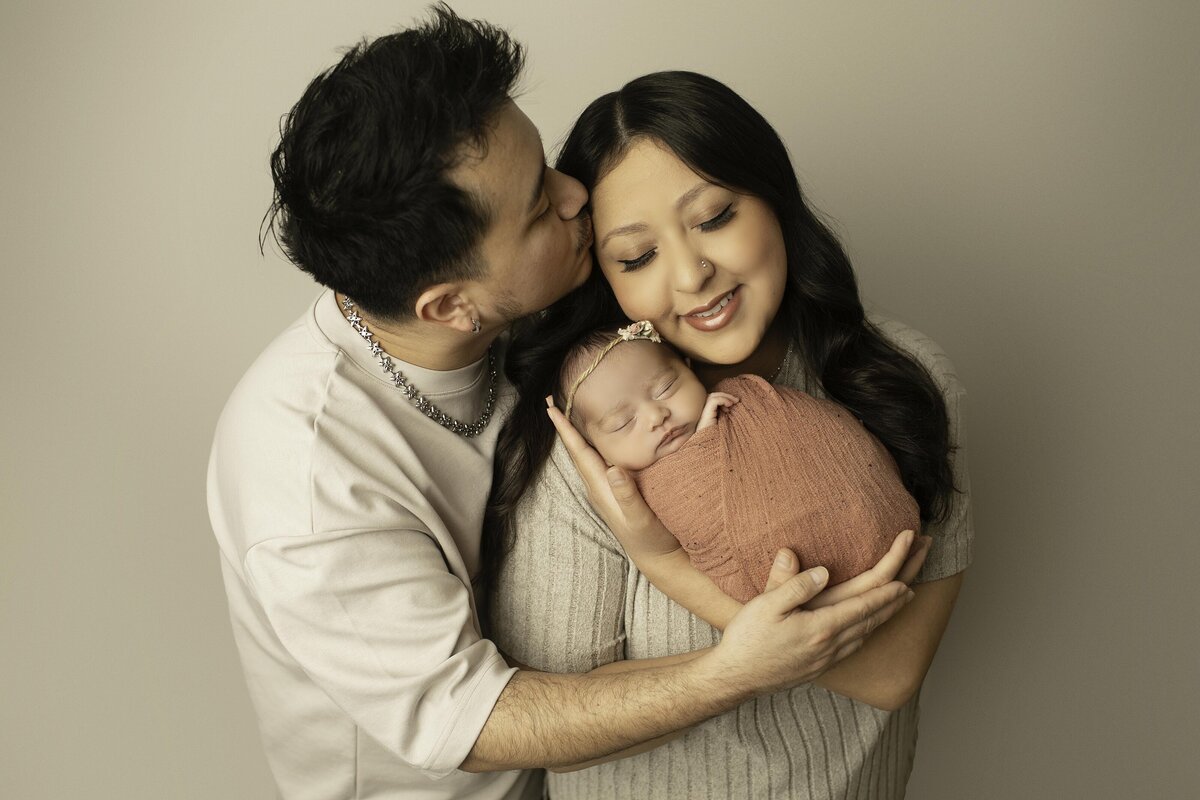 Mom gently cradles her newborn baby while dad lovingly kisses her on the head, capturing a heartfelt and intimate family moment filled with love and warmth