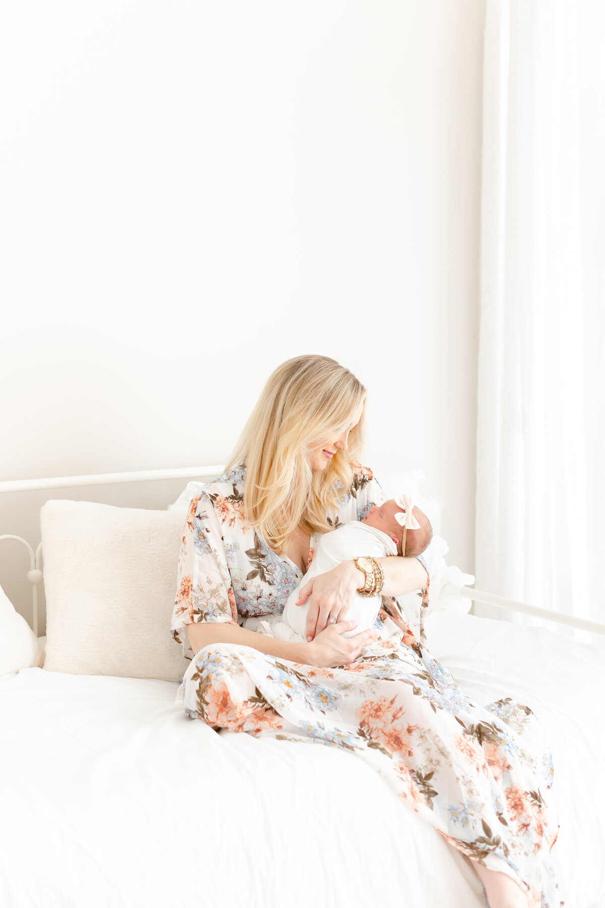 A mother sitting on a beautiful white bed by  a window holding a swaddled baby in her arms by DC Baby Photographer