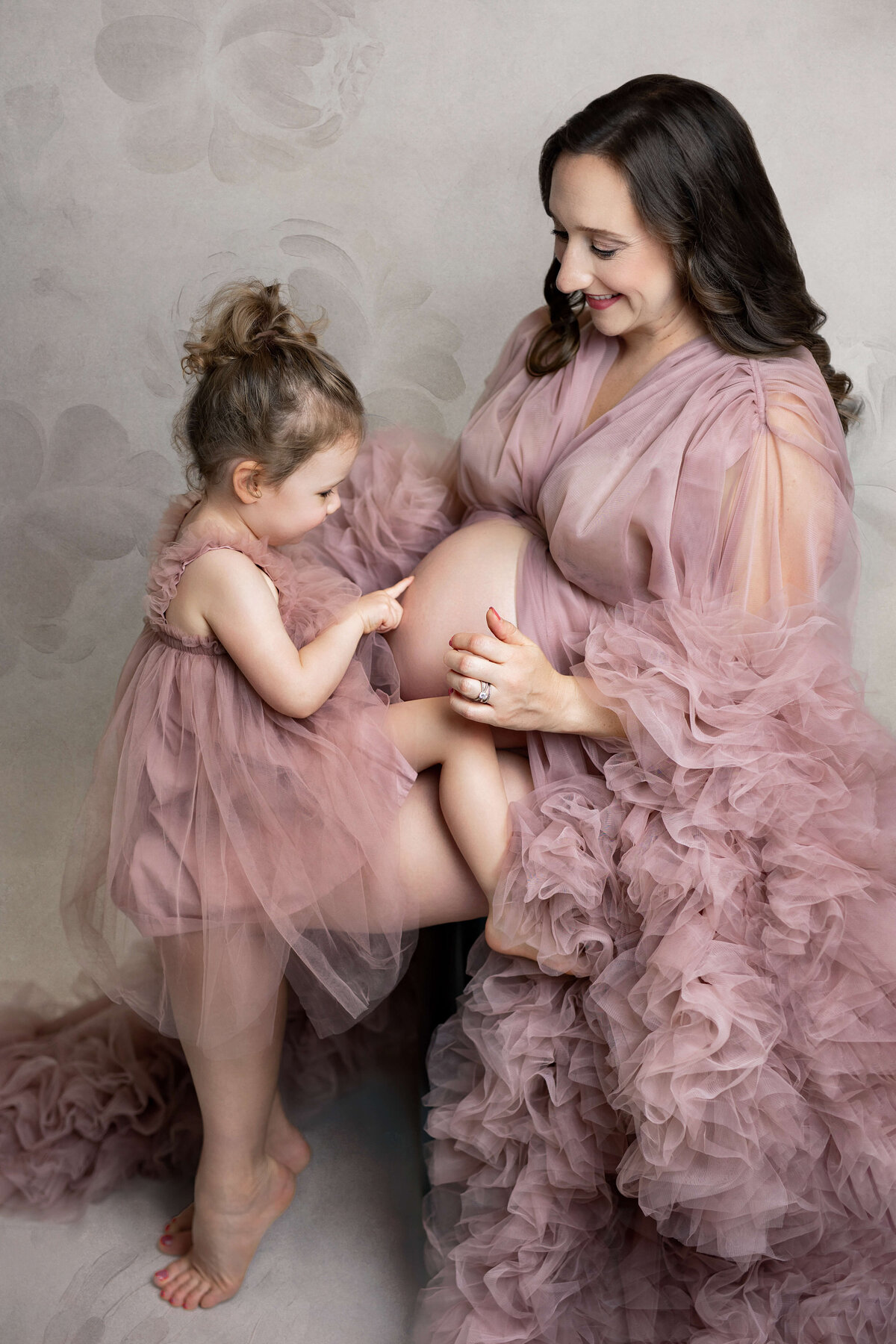 pregnant moter wearing a fluffy pink dress with her little girl sitting on her legs pointing at her pregnant belly