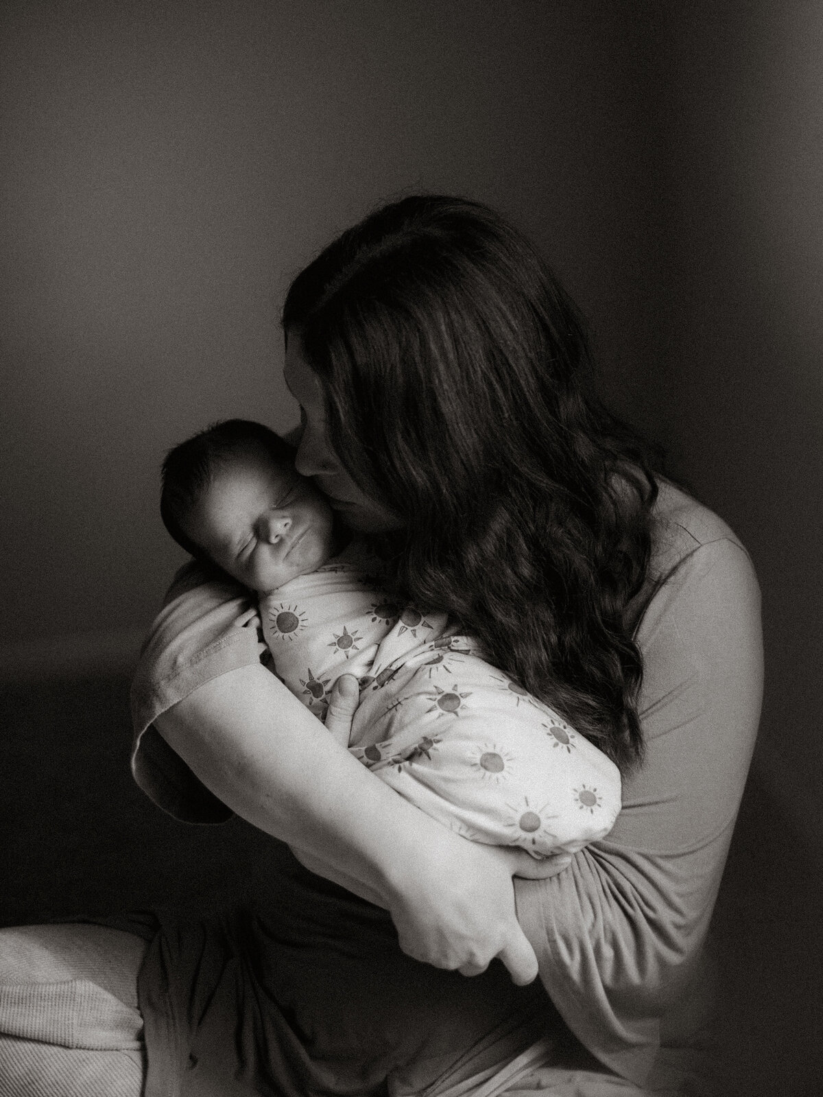 Black and white image of a mother cradling her newborn and gently kissing his cheek by the window during an in-home session in Temecula, CA — a quiet, love-filled moment of early motherhood.