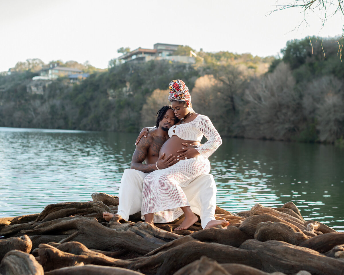 Romantic maternity photo of a couple seated on gnarled tree roots by the lake in Austin, Texas. Mom-to-be wears a white mesh dress and colorful headwrap.