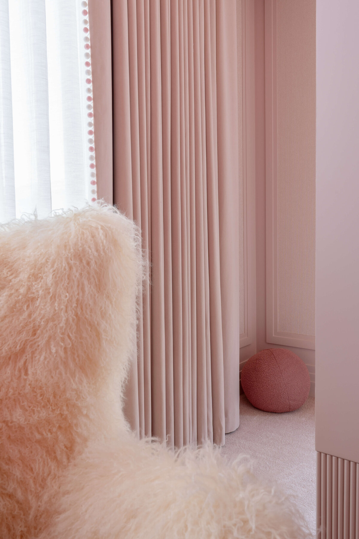 Details of a pink furry armchair in girl's bedroom. Behind the char are soft pink curtains on a large window.